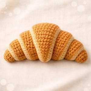 May include: A crocheted croissant in shades of tan and cream, lying on a white surface. The handmade pastry features a detailed, textured stitch pattern, mimicking the look of a freshly baked croissant. The design is a charming, food-themed craft.