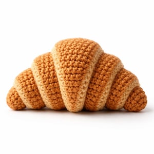 Könnte beinhalten: Ein gehäkelter Croissant in Braun- und Beigetönen. Das handgefertigte Gebäck ist mit strukturierten Stichen versehen, die das Aussehen eines echten Croissants nachahmen. Der Artikel ist vor einem weißen Hintergrund platziert.