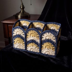 May include: A handcrafted, crocheted organiser in a rectangular form. It displays a repeating pattern of fan-shaped designs in cream, gold, and navy blue. The item is placed on a black surface.