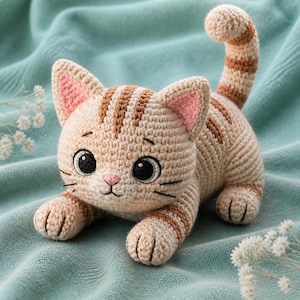 May include: A crocheted cat toy in tan and brown. The cat has large black eyes, pink inner ears, and a pink nose. Brown stripes adorn the head, back, and tail. The cat is lying down on a teal surface.