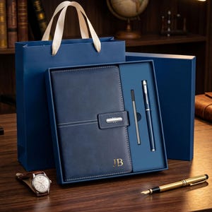 May include: A dark blue leather-bound notebook with a magnetic closure and the gold embossed initials JB, presented in a matching blue gift box with a blue pen and a refill. A blue gift bag and a gold-toned fountain pen sit nearby.