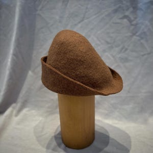 May include: A brown felt hat with a rounded crown and a turned-up brim. The hat is displayed on a wooden hat stand. The hat's color is a warm, earthy tone, and the felt material appears soft and textured.