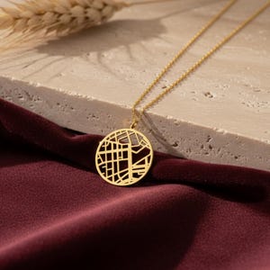 Może przedstawiać: Złoty naszyjnik z okrągłą zawieszką z motywem mapy miasta. Zawieszka wisi na delikatnym łańcuszku. Naszyjnik spoczywa na bordowej aksamitnej powierzchni, z kłosem pszenicy w tle.