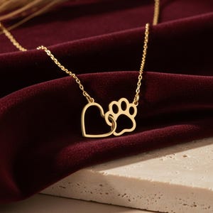 May include: A gold-colored necklace featuring a heart and a paw print pendant. The pendant is connected to a delicate chain. The necklace is displayed on a burgundy velvet surface, with a neutral-colored stone surface in the foreground.