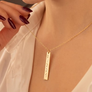 May include: A gold-toned necklace featuring a vertical rectangular pendant. The pendant is engraved with a series of small black dots and dashes arranged in a vertical line, resembling Morse code, suspended from a delicate gold-toned chain.