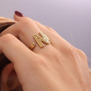May include: A gold-coloured ring featuring the letter "N" in a bold, geometric design. The ring is open at the base and worn on a finger. The ring is a piece of jewellery.