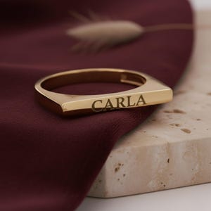 Op de afbeelding: Een goudkleurige ring met de naam "CARLA" gegraveerd. De ring heeft een rechthoekig ontwerp en wordt getoond op een bordeauxrode stof en een stenen oppervlak.