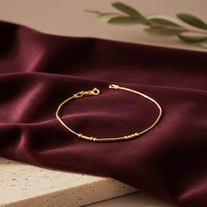 以下が含まれることがあります： 小さなビーズと留め金が付いた繊細なゴールドのブレスレット。深いバーガンディ色のサテン生地の上に置かれています。ブレスレットのシンプルなデザインは、豊かな背景の中で際立っており、上質なジュエリーを連想させます。
