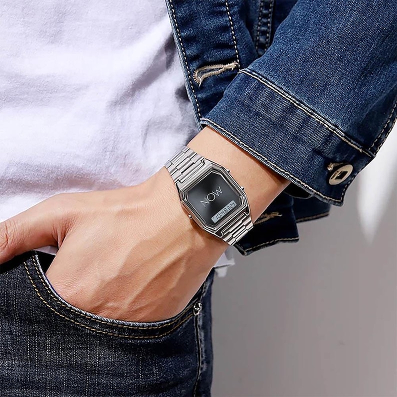 May include: A silver-toned wristwatch with a rectangular black face and a matching metal band. The watch displays the word "NOW" and the time. The watch is worn on a wrist.