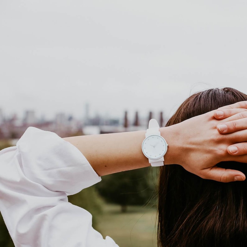 May include: A white wristwatch with a circular face and a white strap. The watch is worn on a wrist, with a white shirt sleeve visible. The background is blurred, showing a cityscape and overcast sky.