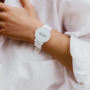 May include: A white wristwatch with a round face and a white band. The watch face displays the word "NOW" in a simple, sans-serif font. The watch is worn on a wrist, with a white shirt in the background.