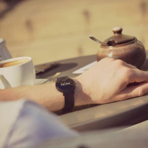 May include: A black smartwatch with the word "NOW" displayed on the screen. The watch has a black band and is worn on a wrist. The image is taken outdoors, with a cup of coffee and a sugar bowl in the background.