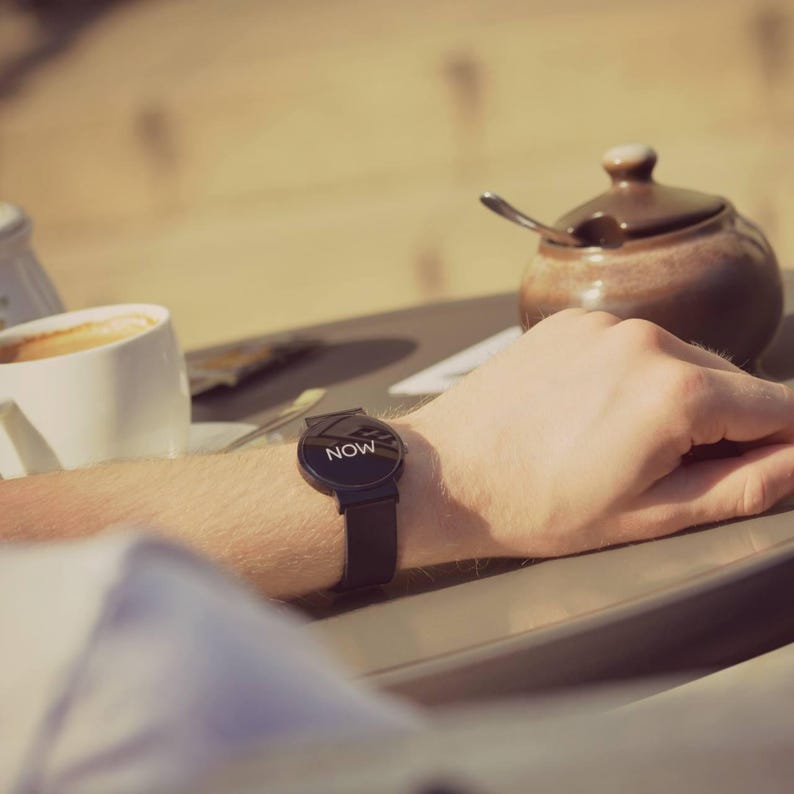 May include: A black smartwatch with the word "NOW" displayed on the screen. The watch has a black band and is worn on a wrist. The image is taken outdoors, with a cup of coffee and a sugar bowl in the background.