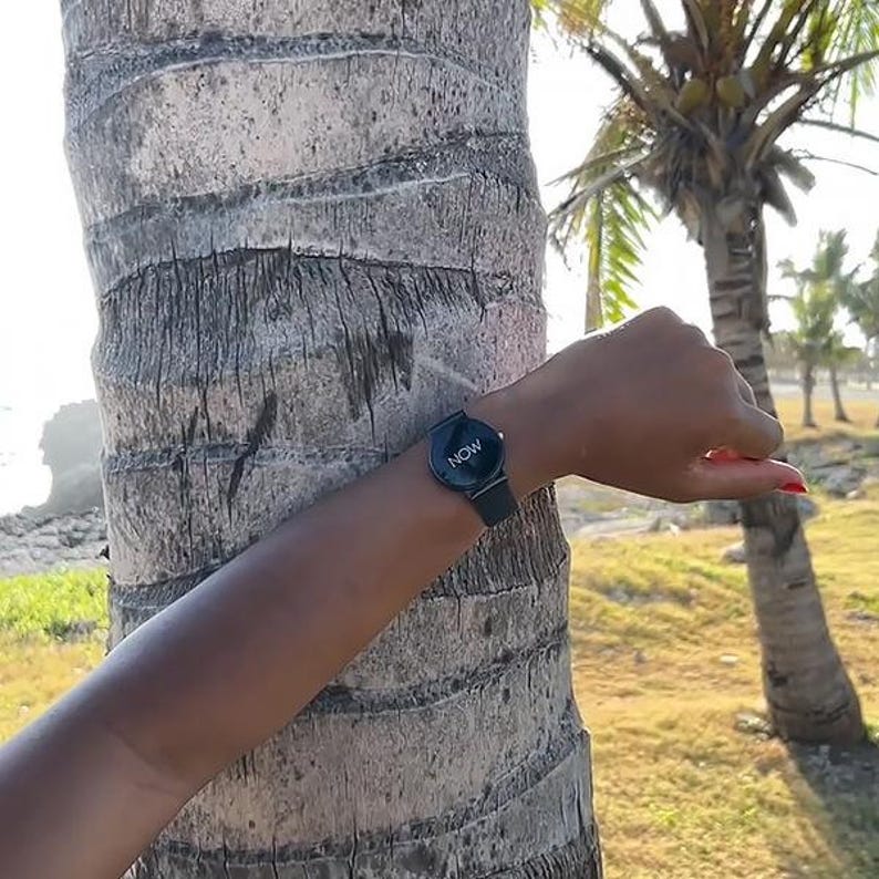 May include: A black wristwatch with the word "NOW" in white lettering on the face. The watch has a black band and is worn on a wrist. The background includes a palm tree and a sunny outdoor setting.
