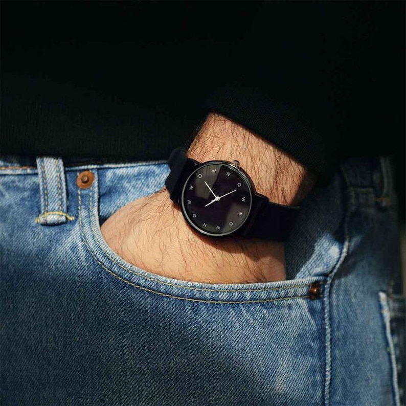 May include: A black wristwatch with a minimalist design. The watch features a black face with white hands and the letters "N" and "W" around the dial. The watch is on a black band.