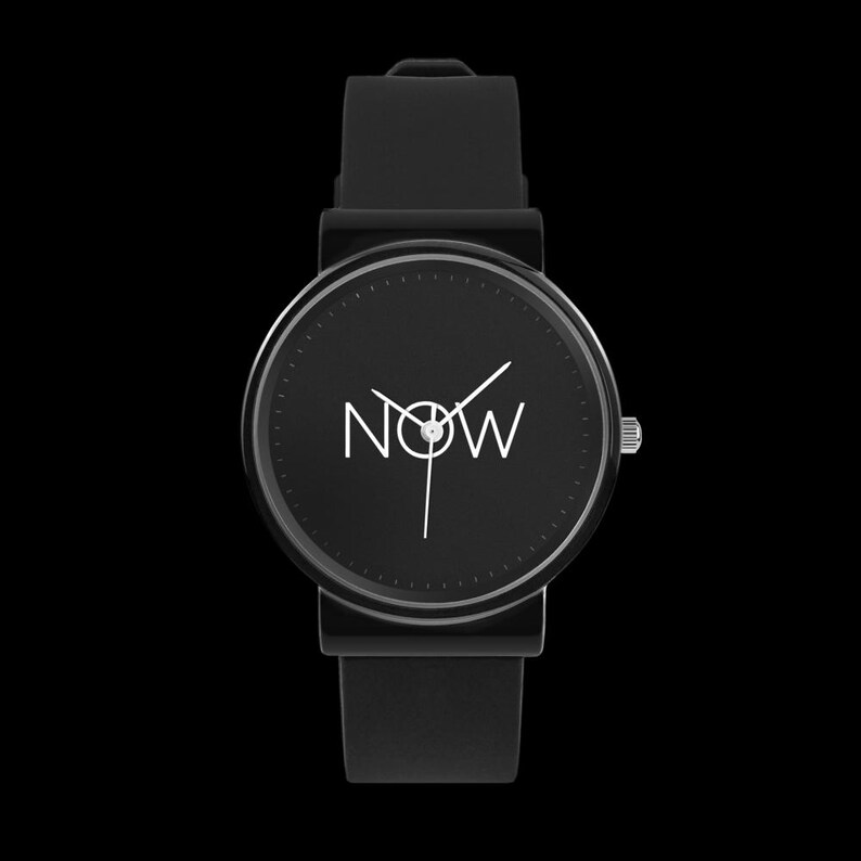 Puede incluir: Reloj de pulsera negro con esfera redonda. La esfera muestra la palabra "NOW" en blanco, con manecillas blancas que indican la hora. El reloj tiene una correa negra y una corona plateada. El fondo es negro liso.