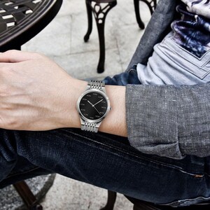 May include: A silver wristwatch with a black face and silver band. The watch face has the word "NOW" in a circle around the center. The watch is on a wrist, with a denim-clad arm and a gray long-sleeved shirt visible.