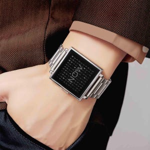 Peut inclure: Montre-bracelet argent&eacute;e avec un cadran carr&eacute; noir affichant le mot "NOW" en grandes lettres. Le cadran est entour&eacute; d'une grille de petites lettres. Le bracelet est en m&eacute;tal argent&eacute;.