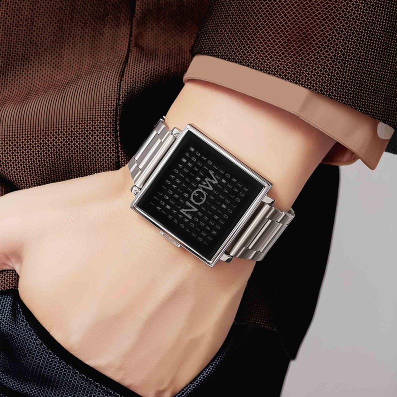 Peut inclure: Montre-bracelet argent&eacute;e avec un cadran carr&eacute; noir affichant le mot "NOW" en grandes lettres. Le cadran est entour&eacute; d'une grille de petites lettres. Le bracelet est en m&eacute;tal argent&eacute;.