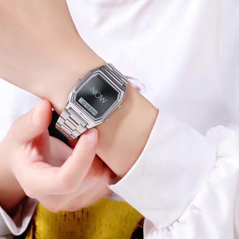 May include: A silver-toned digital watch with a rectangular face and matching band. The watch face displays the word "NOW" and the time. The watch is worn on a wrist, with a white shirt sleeve in the background.