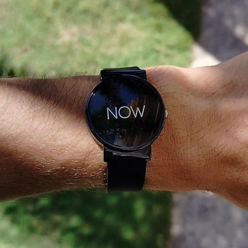 May include: Black smartwatch with a round face displaying the word "NOW" in white. The watch has a black band and is worn on a wrist. The background is blurred green.