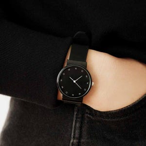 May include: A minimalist black wristwatch with a round face and a black band. The watch face features white hands and the letters "N", "O", and "W" in place of numbers. The watch is worn on a wrist, with a black sleeve and dark denim jeans in the background.