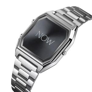 Puede incluir: Reloj de pulsera plateado con una esfera rectangular gris oscuro y una correa de metal a juego. La esfera del reloj muestra la palabra "NOW" en plata. El reloj tiene un diseño moderno y minimalista.
