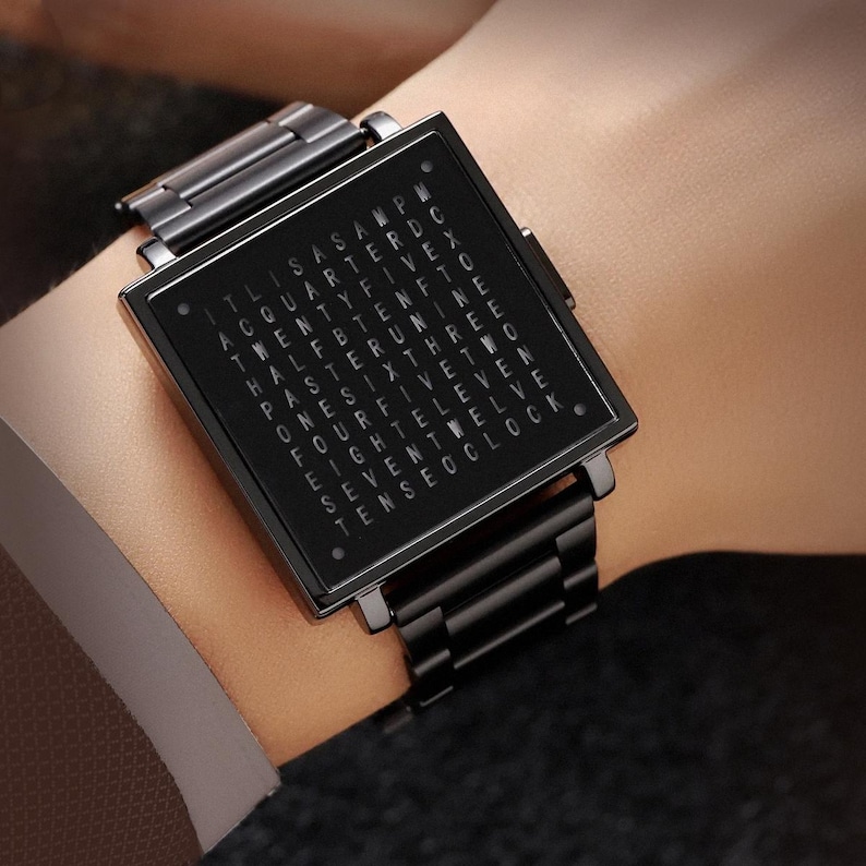 May include: A black square watch with a metal band is displayed on a wrist. The watch face has a grid of words indicating the time. The watch is a dark, sleek design.