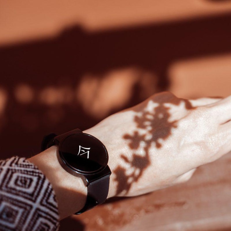 Puede incluir: Un reloj inteligente negro con una esfera redonda y una correa negra se lleva en la mu&ntilde;eca. La esfera del reloj muestra un s&iacute;mbolo blanco. El fondo es de un color marr&oacute;n rojizo c&aacute;lido, con sombras de hojas en la mu&ntilde;eca.