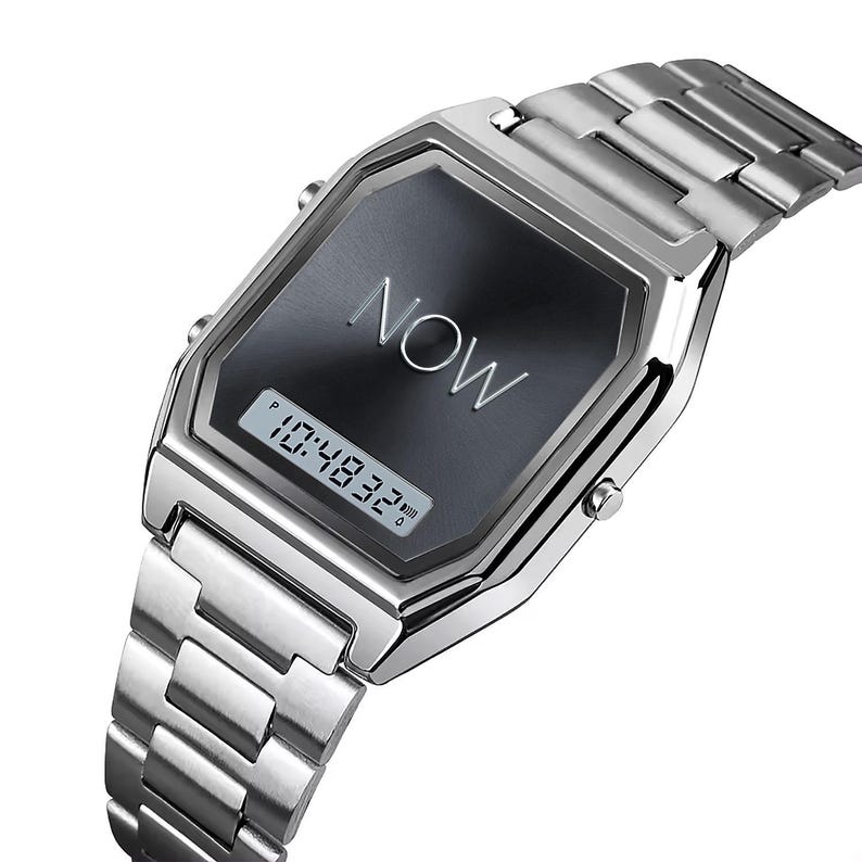 May include: A silver-toned digital watch with a rectangular face. The watch face is black with the word "NOW" in silver lettering. The digital display shows the time as 10:48:32. The watch has a metal band.