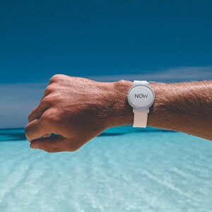 May include: A white wristwatch with a round face and a white band. The watch face displays the word "NOW" in a simple, sans-serif font. The watch is worn on a wrist against a backdrop of blue water and sky.