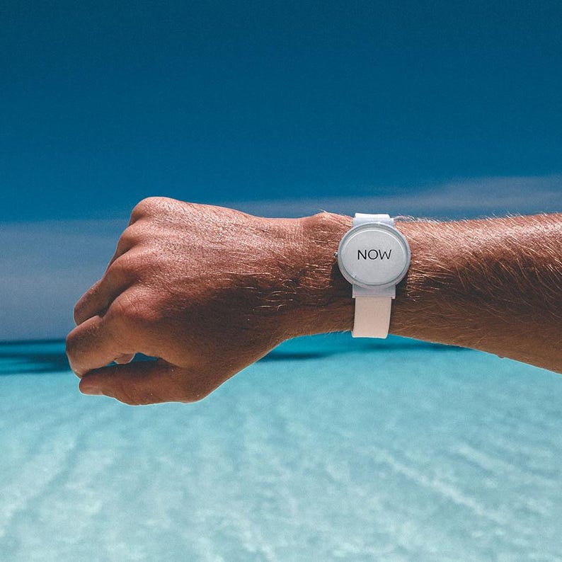 May include: A white wristwatch with a round face and a white band. The watch face displays the word "NOW" in a simple, sans-serif font. The watch is worn on a wrist against a backdrop of blue water and sky.