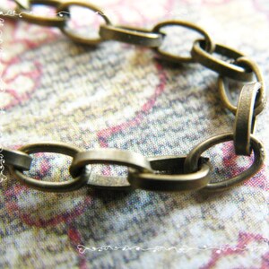 May include: Close-up of a section of a bronze-colored chain. The oval links are interlocked, creating a textured pattern. The chain is set against a soft, patterned background with hints of pink and cream.