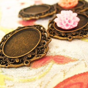 Puede incluir: Marcos de colgantes ovalados de color bronce ornamentados, con bordes de filigrana decorativos. Algunos marcos contienen cabujones de flores rosas y melocotón. Los marcos están sobre una tela con estampado floral.
