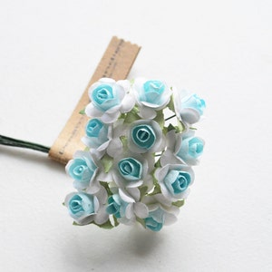 以下が含まれることがあります： 白い花びらと水色の中心を持つ小さな紙のバラの束。花は集められ、茶色の紙バンドでまとめられています。緑色の茎が花の根元から伸びています。