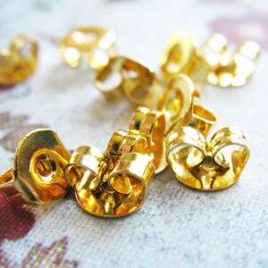 May include: A close-up of a group of gold-colored earring backs. The backs are made of metal and have a curved shape. They are designed to hold earrings in place.