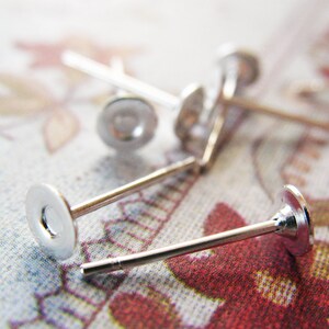 May include: Close-up of several silver-colored earring posts with flat, round backs. The posts are thin and straight, with a small, circular disc at the end. The earrings are scattered on a floral patterned surface.