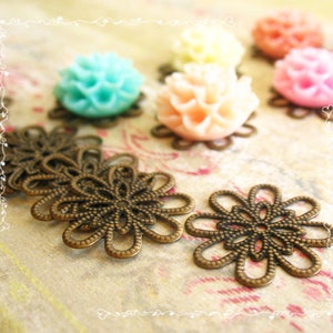 May include: Close-up of bronze-colored filigree flower embellishments with a detailed openwork design. Several colorful resin flower cabochons in shades of pink, peach, yellow, and turquoise are also visible, suggesting a craft supply theme.