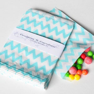 May include: A stack of white paper bags featuring a light blue chevron pattern, secured with a white paper band printed with the text Dreams & Candies Lovely goods. One open bag contains a collection of small, round, multi-colored candies.