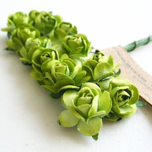 以下が含まれることがあります： 一列に並んだ小さなライムグリーンの造花のバラの束。それぞれの花はマットな質感の花びらが重なり合っています。バラは細い緑色のワイヤーの茎に取り付けられており、それらがまとめられ、茶色の紙の帯で巻かれています。