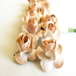 以下が含まれることがあります： 柔らかいベージュとタンのカラーパレットで構成された、小さな造花のバラの束。それぞれの花は、層になった丸みを帯びた花びらでできており、根元は少し明るい色合いになっています。背面からは細い緑色のワイヤーの茎が伸びています。