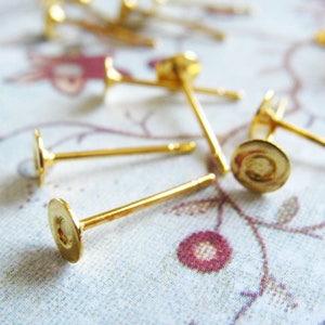 May include: Close-up of several gold-colored earring posts with flat, round pads. The posts are thin and straight, with a small, flat disc at the end for attaching earrings. The background is a soft, floral pattern.
