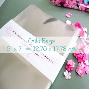 May include: A stack of clear cellophane bags with a white label that reads "Dreams & Lovely goods & Indies Cello Bags 5" x 7" = 12.70 x 17.78 cm". Pink confetti is scattered around the bags.
