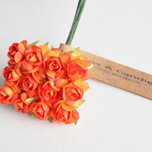 May include: A cluster of small, vibrant orange paper roses attached to thin green wire stems. A rectangular brown paper tag is attached to the stems, featuring the partially visible printed text "ns & Candies". The roses have layered petals.
