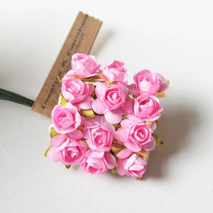 以下が含まれることがあります： 層状の花びらと基部に緑色の紙製のがくを持つ、12個の小さな明るいピンク色の紙のバラの束。バラは細い濃い緑色のワイヤーの茎に取り付けられており、小さな茶色の長方形のタグがワイヤーに付いています。