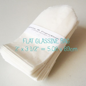 以下が含まれることがあります： 「Jams & Candies」および「FLAT GLASSINE BAG 2" x 3 1/2" = 5.08 x 8.89cm」と書かれた紙帯でまとめられた、半透明の白い平らなグラシン紙袋の束。袋は長方形で、上端が丸みを帯びた形状をしています。
