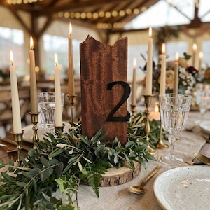 May include: A wedding table setting with a wooden table number displaying the number "2". The table is adorned with foliage, lit candles in brass holders, and fine glassware. The scene evokes a sense of celebration and elegance.