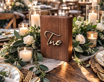 May include: A rustic wooden table setting with a centerpiece featuring a wooden block with the number "Two" in gold script. The table is decorated with white roses, greenery, candles, and gold-toned cutlery, creating an elegant ambiance.