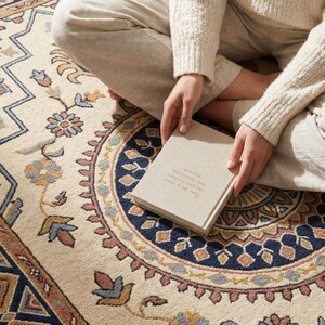 May include: Cream-coloured area rug with a detailed floral and geometric pattern in shades of blue, brown, and peach. A book is held in the lap, with text visible. The rug's design features a central medallion and border.
