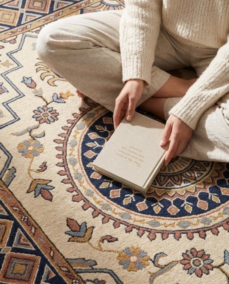 May include: Cream-coloured area rug with a detailed floral and geometric pattern in shades of blue, brown, and peach. A book is held in the lap, with text visible. The rug's design features a central medallion and border.